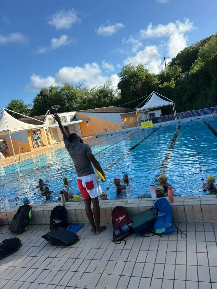 Un entraîneur, bras levé, donne les consignes au bord du bassin, les sacs des nageurs alignés derrière lui