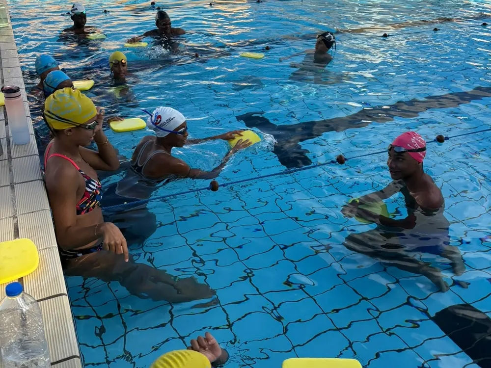 Des nageuses en bonnets jaune, blanc et rose font un exercice avec une planche contre le bord du bassin